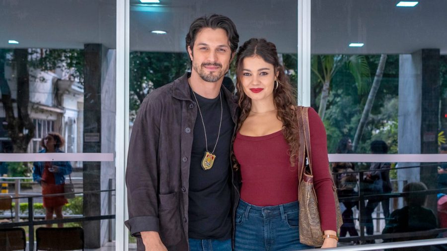 Uma fotografia em plano médio dos atores Sophie Charlotte e Romulo Estrela, como Gerluce e Paulinho, posando lado a lado na frente de portas de vidro grandes e modernas. Paulinho (Romulo Estrela, à esquerda) usa uma jaqueta de couro marrom aberta sobre uma camiseta preta e um distintivo de polícia em uma corrente em volta do pescoço. Gerluce (Sophie Charlotte, à direita) veste um top de mangas compridas vermelho escuro de gola canoa e jeans de cintura alta azuis, carregando uma bolsa de ombro de couro marrom texturizada. Paulinho tem o braço terno ao redor da cintura dela e ambos estão sorrindo para a câmera. O fundo de vidro reflete árvores verdes tropicais e reflexos desfocados de outras pessoas do lado de fora do prédio. A iluminação é suave e uniforme