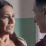 Três Graças: Resumo dos Capítulos, Elenco e Spoilers da Novela das 21h 2 Uma fotografia em plano médio dos atores Dira Paes e Enrique Diaz, como Lígia e Albérico, posando lado a lado em um interior de Três Graças. Lígia (à direita, com cabelos longos escuros ondulados e blusa branca com laço no pescoço) sorri terna e apaixonadamente para Albérico (à esquerda, com cabelos curtos grisalhos ondulados e jaqueta de linho bege), que sorri de volta com igual afeto. Eles estão de mãos dadas e trocam olhares íntimos. O fundo é desfocado, mostrando cortinas de linho claro e marrom e uma janela. A iluminação é suave e natural.