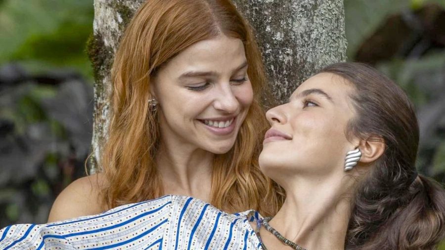 Fotografia de cena carinhosa entre as atrizes Alanis Guillen (ruiva, como Lorena Ferette) e Gabriela Medvedovsky (morena, como Juquinha ou Eduarda Fragoso), que interpretam o casal Lorena e Juquinha na novela das nove da TV Globo, "Três Graças". Lorena (Alanis Guillen) tem cabelos ruivos longos e sorri ternamente para Juquinha (Gabriela Medvedovsky), que está recostada em seu colo em um ambiente de jardim ou área verde. Juquinha tem cabelos morenos em um rabo de cavalo e olha afetuosamente para Lorena, usando um brinco de prata em forma de concha e colar de miçangas. O momento captura a intimidade e o romance genuíno entre as personagens que desafiam barreiras na trama paulistana.